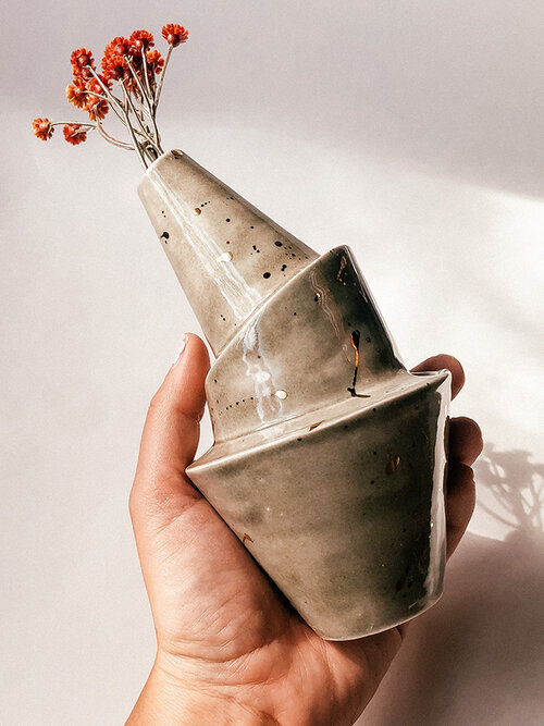 Hand holding Wandering Bud bubbler in charcoal with gold splatters. Small red, orange flowers coming out of top.